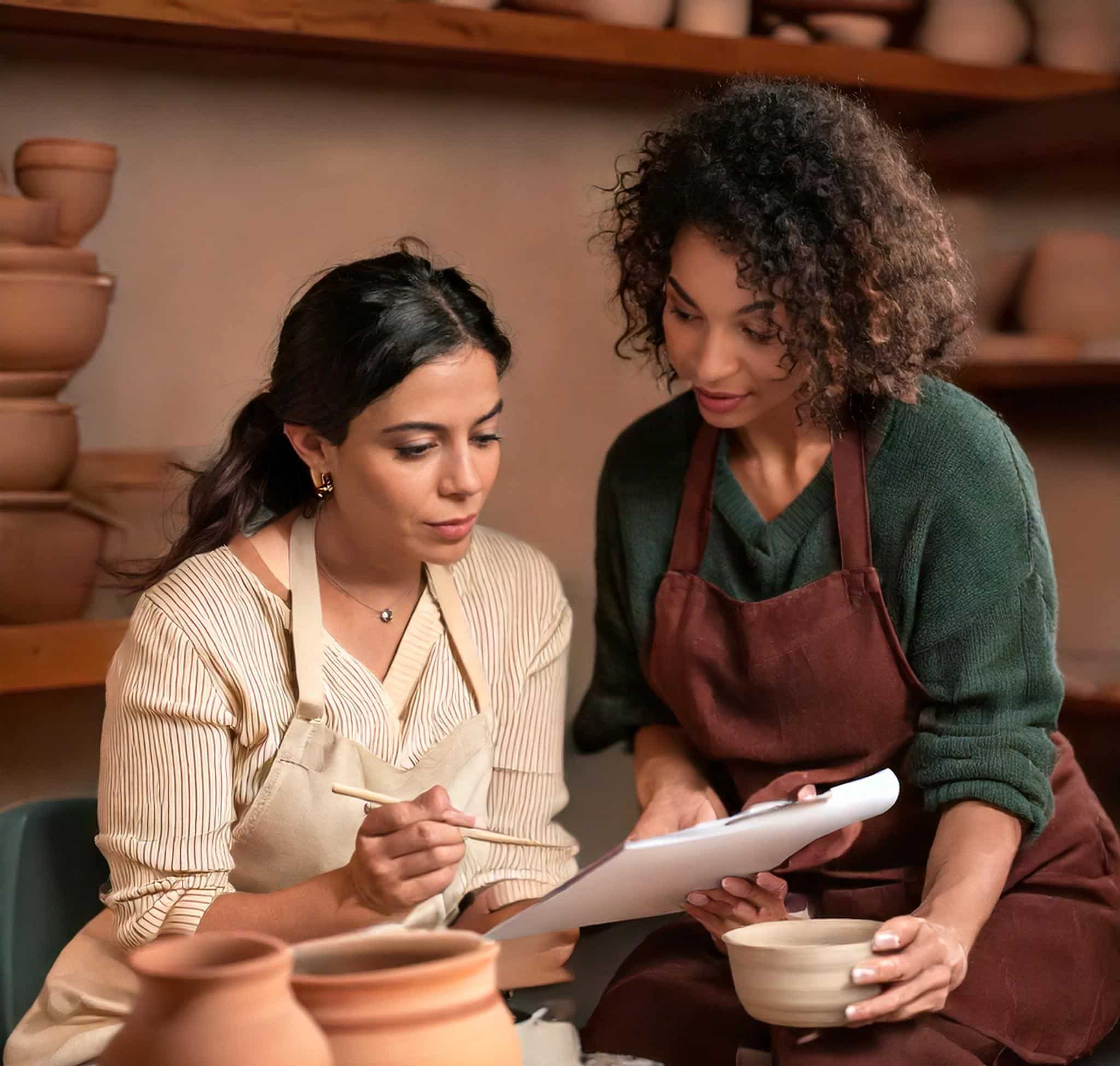 Deux femmes de 40 ans brune sont dans un atelier de poterie. L'une d'elle écrit sur une feuille blanche.